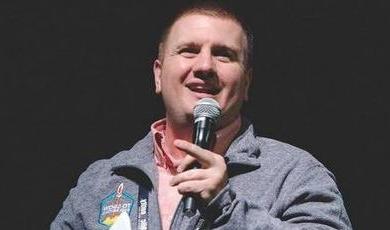 Mickey Howard stands in front of a black background holding a microphone