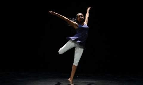 female dancer on stage wearing light gray cropped leggings and a royal purple tunic top standing on one pointed toe with her arms up and outstretched and her left leg bending behind her in a dance