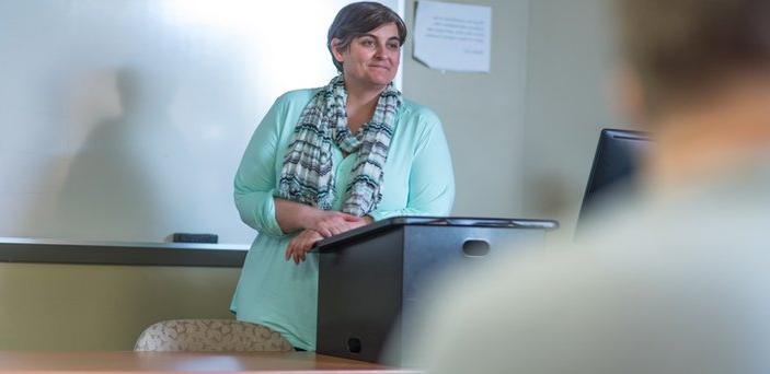 A professor teaching a class and smiling at a podium in front of a white board