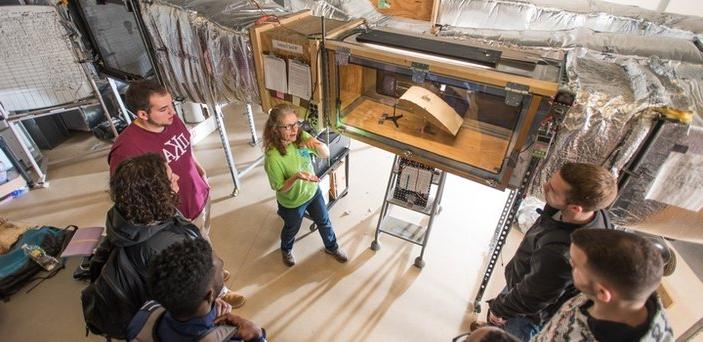 A professor with a group of students in front of the wind tunnel